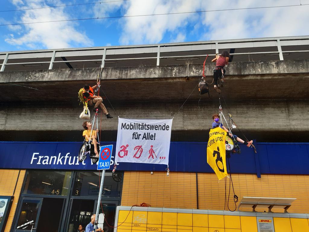 Kletteraktion an der Fassade der Bahnhofes Frankfurt West, 4 menschen, 2 in Rollstuhl, alle in Seile gesichert und Banner zwischen ihnen  es steht Mobilitätswende für Alle