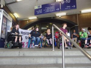Viele Leute im Rollstuhl sitzen unter das Schild fuer Gleis 9-10 am Messe Deutz. Im Vordergrund ist eine Steile Treppe.
