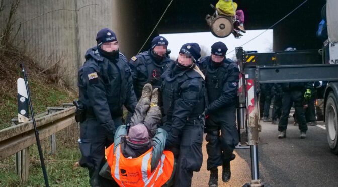 Lützerath, eine Person wird von der Polizei weg getragen. Im Hintergrund eine kletternde Person im Rollstuhl an einer Brücke im Seil