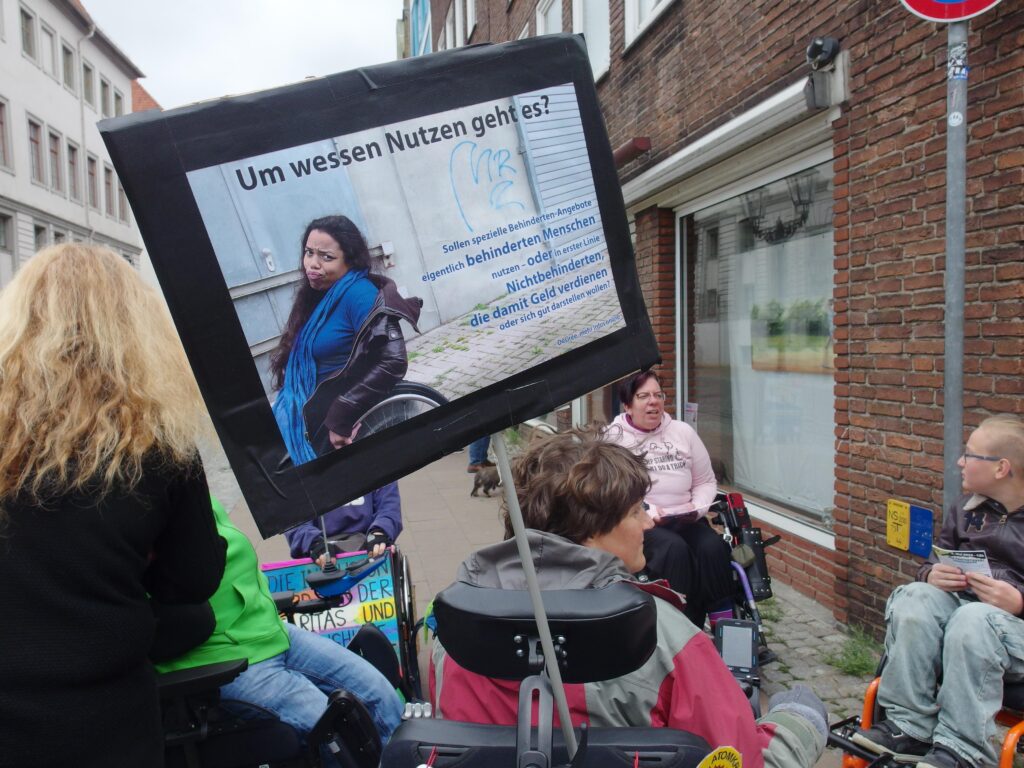 Demonstrierende, ein Plakat: Eine junge, afro-deutsche Frau fährt mit ihrem Rollstuhl seitlich aus dem Bild heraus. Sie guckt verärgert in die Kamera und zieht eine Grimasse.
Ihr Text: Um wessen Nutzen geht es? Sollen spezielle Behinderten-Angebote eigentlich behinderten Menschen nutzen – oder in erster Linie Nichtbehinderten, die damit Geld verdienen oder sich gut darstellen wollen?
Zur Person: Désirée, mehr Infos online