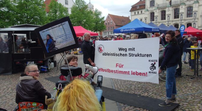 Menschen mit und ohne Rollstuhl mit Megafon, Banner "Weg mit ableitischen Strukturen. Für selbstbestimmt leben! im Hintergrund ein Stand der Lebenshilfe