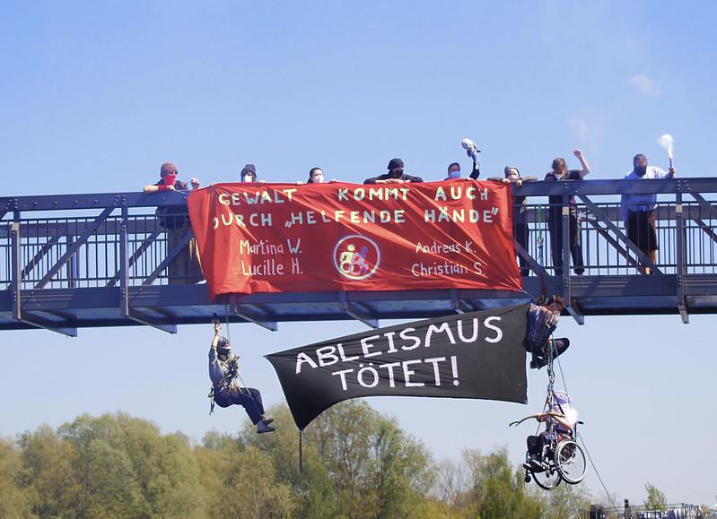 Aktivist:innen hängen unter einer Brücke an Seilen; eine Person im Rollstuhl ist ebenfalls gesichert. Oben auf der Brücke halten weitere Aktivist:innen ein rotes Transparent mit der Aufschrift „Gewalt kommt auch durch ‚helfende Hände‘“, darunter stehen die Namen Martina W., Lucille H., Andreas K. und Christian S. Unterhalb hängt ein schwarzes Banner mit der Aufschrift „Ableismus tötet!“. Einige Personen halten Rauchfackeln. (Foto: ProtestEyes PE)