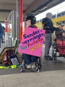 1 Mensch im Rollstuhl spricht ins Megafon, 1 mensch im Rollstuhl trägt ein Plakat " Sondervermögen für Aufzüge jetzt