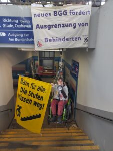 1 Menschen hängt im Rollstuhlsitzend in Seilen in einem Treppenhaus am Bahnhof. zwei Banner: Bahn für alle die Stufen müssen weg und neues BGG fördert Ausgrenzung von Behinderten