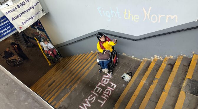 Blick auf das Treppenhaus am Bahnhof Buxtehude. Hintergrund, eine Person im Rollstuhl in der Luft im Seil gesichert, im Vordergrund 1 Mensch mit Rollstuhl. und Kreisde Spruch "Fight Ableism" auf der Treppe, an der Wand "Fight the Norm"