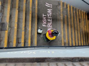 1 Mensch sitz auf der Treppe neben dem Kreidespruch "Fight Ableism"
