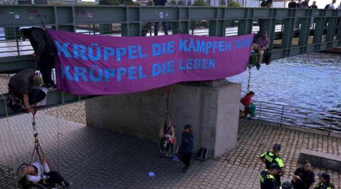 großes lila Banner "Krüppel die kämpfen sind Krüppel die leben" an einer Brücke 6 menschen klettern, 3 davon in seilen im rollstuhl sitzend gesichert. im Hintergrund ist der Reichtstag zu sehen
