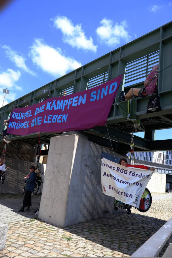 Banner an einer Brücke. "Krüppel die kämpfen sind Krüppel die leben" darunter menschen die im Rollstuhl sitzend klettern, in seilen gesichert sind. rechts hält die Person ein weiteres Banner in der Hand " neues BGG fördert Ausgrenzung von Behinderten"