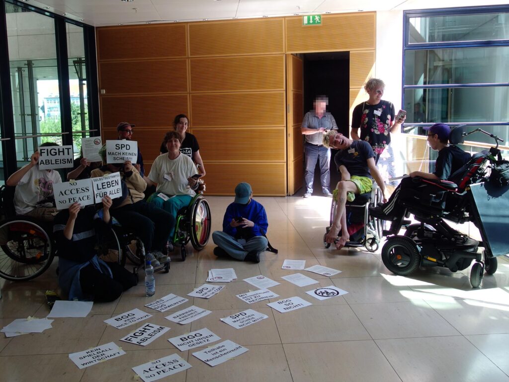 Gruppe von Aktivist*innen bei der Besetzung mit zahlreichen Schildern "fight ableism, no access no peace, SPD mach keinen scheiß, selbtbestimmtes Leben für alle, kein Speilball der Wirtschaft" etc.