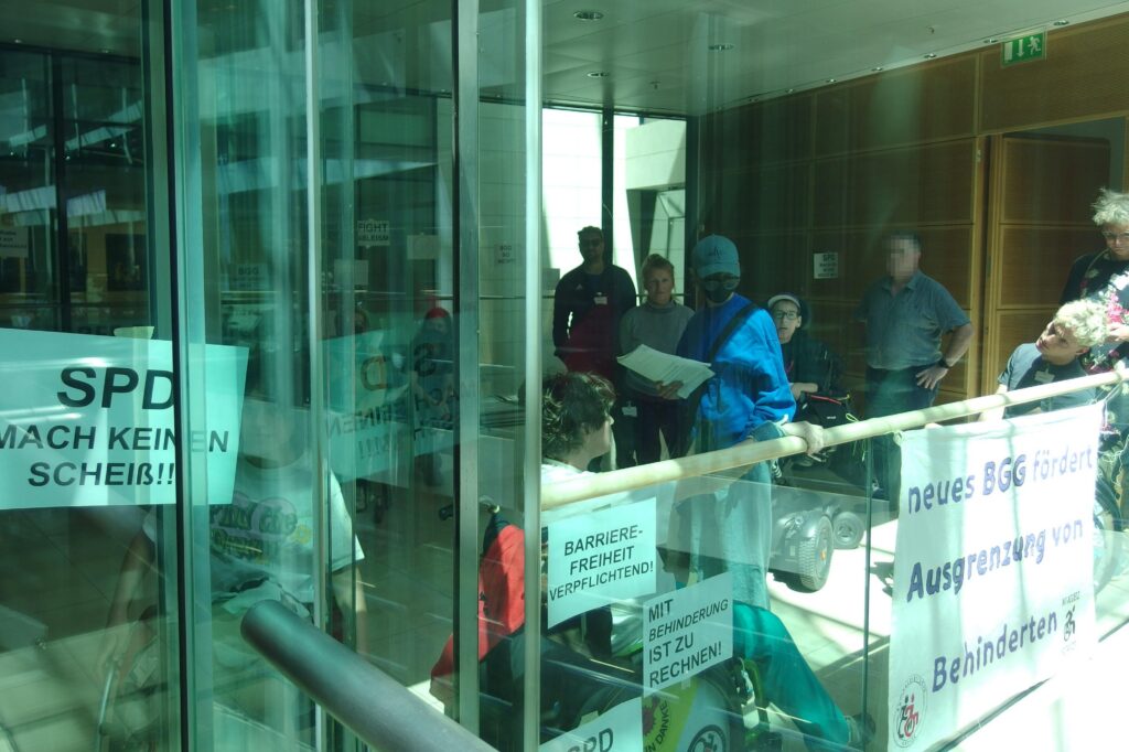 Besetzung vom Glasaufzug aus fotografiert. Banner an der Ballustrade " neues BGG fördert Ausgrenzung von Behinderten" und viele Zetteln " SPD mach keinen Scheiß", "Barrierefreiheit verpflichtend, Mit Behinderung ist zu rechnen"