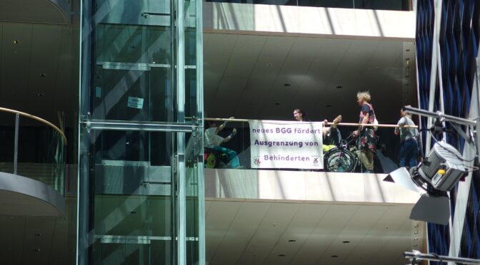 menschen sind an der Ballustrade aus Glas von einem Stockwerk zu sehen, einige haben einen Rollstuhl. Sie haben ein Banner an der Ballustrade angebracht "neues BGG fördert Ausgrenzung von Behinderten". und links beim Glasaufzug ein Schild "SPD mach keinen Scheiß"